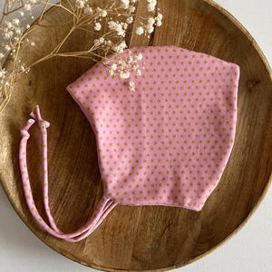 Può includere: Un cappellino rosa per neonati con un motivo a stelle dorate. Il cappellino ha lunghi lacci rosa abbinati. Il cappellino è esposto su un piatto di legno con fiori bianchi secchi.