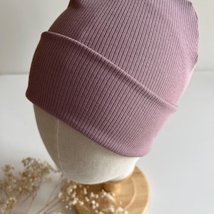 Può includere: Un berretto lavorato a coste di colore malva. Il cappello è esposto su una testa di manichino con base in legno. Il bordo ripiegato e la trama a coste del cappello sono ben visibili. Lo sfondo è bianco neutro.