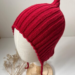 May include: A vibrant red knit hat with a unique, pointed top and ribbed texture. The hat features ear flaps with attached ties and a scalloped edge. This handmade hat is displayed on a mannequin head.