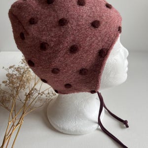 Può includere: Un berretto per neonati in lana rosa con pois marroni e un laccio marrone.