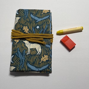 Op de afbeelding: Een stoffen oproletui met een donkerblauw patroon van wolven, vogels, eikenbladeren en sterren, vastgezet met een mosterdgeel koord. Naast het etui liggen een geel waskrijtje met de tekst "Nr. 5 Zitronengelb WACHSMALSTIFT" en een klein rood blokje.