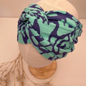 Könnte beinhalten: Ein geknotetes Stirnband mit einem abstrakten Muster in Türkis und Marineblau wird auf einem Schaufensterkopf präsentiert. Der Stoff ist weich und scheint ein leichtes Material zu sein. Das Stirnband ist ein stilvolles Accessoire.