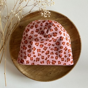 Op de afbeelding: Een roze beanie met een luipaardprint in bruin en oranje. De hoed ligt op een houten bord, met gedroogde bloemen op de achtergrond. Een modieus accessoire.