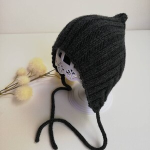 Könnte beinhalten: Eine schwarze gestrickte Baby-Mütze mit spitzer Spitze und langen Bändern. Die Mütze ist aus einem weichen, warmen Material gefertigt und perfekt, um den Kopf eines Babys im Winter warm zu halten.
