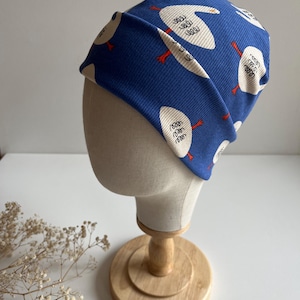 Op de afbeelding: Een blauwe beanie met een wit ganzenpatroon en rode accenten wordt getoond op een mannequinhoofd. De hoed is gemaakt van een geribbelde stof. Het mannequinhoofd staat op een houten standaard. Een klein label met het woord "hello" is zichtbaar.