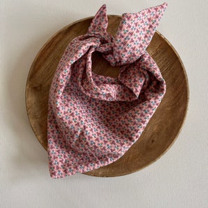 Può includere: Un bandana rosa con un motivo floreale, annodato e mostrato su una superficie di legno.