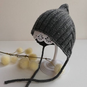 Könnte beinhalten: Eine graue gestrickte Baby-Mütze mit spitzer Spitze und langen Bändern. Die Mütze wird auf einem weißen Ständer mit einer Spitzenverzierung präsentiert.