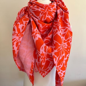 Puede incluir: Una bufanda vibrante con un estampado floral en tonos naranja y rosa. La bufanda está drapeada sobre una cabeza de maniquí blanca, mostrando su diseño y textura. La tela parece ligera y fluida, perfecta para añadir un toque de color a cualquier atuendo.