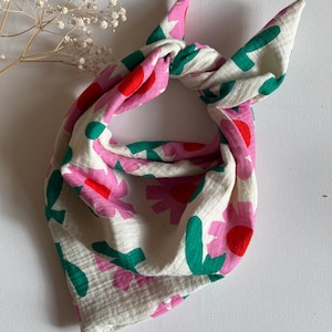 Könnte beinhalten: Ein geknotetes Halstuch mit floralem Muster. Der Schal hat einen cremefarbenen Hintergrund mit rosa, roten und grünen Blumenformen. Der Stoff ist weich und strukturiert.