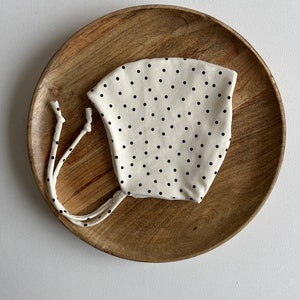 Pode incluir: Um gorro branco com bolinhas pretas e um cordão para amarrar. O gorro está sobre um prato de madeira.