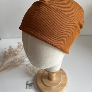 Può includere: Un berretto a coste di colore arancione ruggine. Il cappello è esposto su una testa di manichino con un supporto in legno. Piccole etichette con testo sono visibili vicino alla base.
