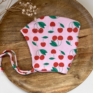 Pode incluir: Um gorro de bebé rosa claro com um padrão repetido de cerejas vermelhas e folhas verdes. O gorro tem um fecho de laço e é feito de tecido macio. As cerejas têm rostos sorridentes. O gorro está numa bandeja de madeira.