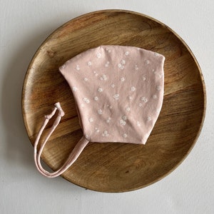 Puede incluir: Un gorro de bebé rosa con un estampado floral blanco y lazos. El gorro está sobre una superficie de madera.