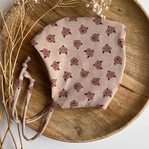 Può includere: Un berretto per neonato rosa chiaro con un motivo di piccole facce di orso marroni. Il berretto ha due lunghi lacci che possono essere legati sotto il mento.