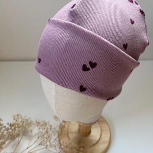 Puede incluir: Un gorro lila con textura acanalada y patrones de corazón morado oscuro. El gorro se exhibe en una cabeza de maniquí con base de madera. Un accesorio de moda.