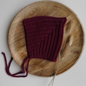 Könnte beinhalten: Eine bordeauxfarbene, gestrickte Babyhaube mit Bindebändern. Die Haube ist aus Rippstrick gefertigt und wird auf einem Holztablett präsentiert. Die Haube hat eine tiefrote Farbe und eine quadratische Form.