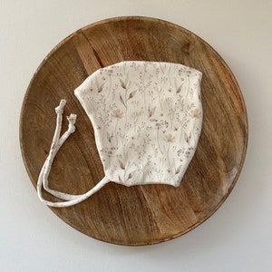 Puede incluir: Un gorro blanco con un patrón floral en tonos marrones y beige. El gorro tiene dos lazos largos que se pueden atar debajo de la barbilla.