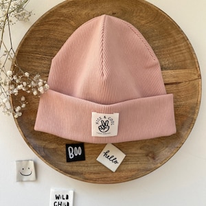 Puede incluir: Un gorro de color rosa empolvado con canalé y un dobladillo. Una etiqueta blanca con el texto "NICE & COOL" y un signo de la paz está cosida en la parte delantera. También hay pequeñas etiquetas con las palabras "boo", "hello" y "WILD CHILD".