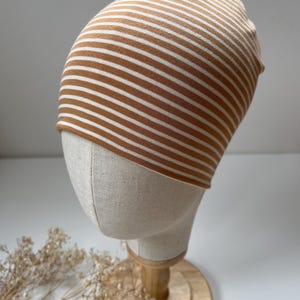 May include: A brown and white striped beanie hat displayed on a mannequin head. The hat is made of a soft material and features horizontal stripes. The mannequin head is on a wooden stand, and the background is a neutral color.