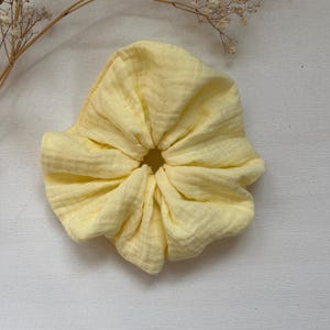 Könnte beinhalten: Ein hellgelbes Scrunchie aus weichem, strukturiertem Stoff. Das Haaraccessoire ist in der Mitte gerafft, wodurch ein gerüschter Effekt entsteht. Das Scrunchie ist einfarbig und scheint aus einem leichten Material zu bestehen.