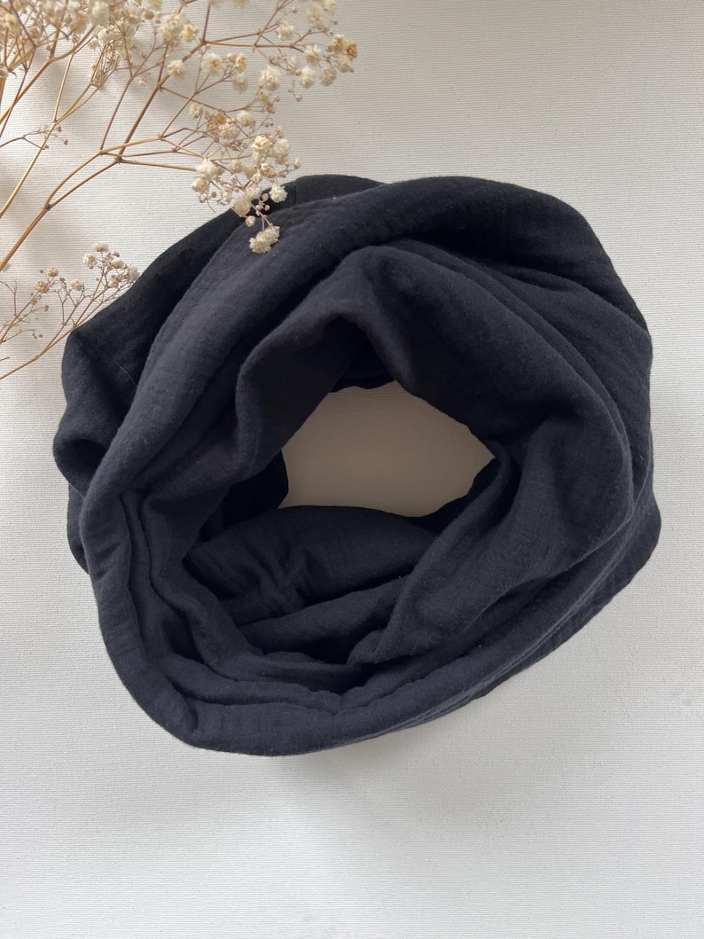 Peut inclure: Une &eacute;charpe noire en tissu doux. L'&eacute;charpe est enroul&eacute;e en cercle et est pos&eacute;e &agrave; plat sur une surface blanche.