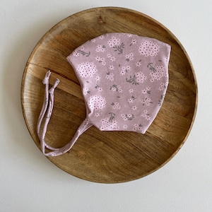 Può includere: Un berretto di stoffa rosa con un motivo floreale. Il berretto ha due lunghi lacci.