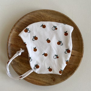 Può includere: Cappellino bianco per neonato con stampa di frutta marrone e bianca. Il cappellino ha un laccio sul retro.