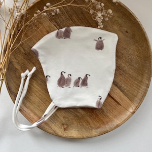Pode incluir: Um gorro branco para bebê com ilustrações de pinguins marrons. O gorro tem duas ataduras brancas que podem ser amarradas sob o queixo.