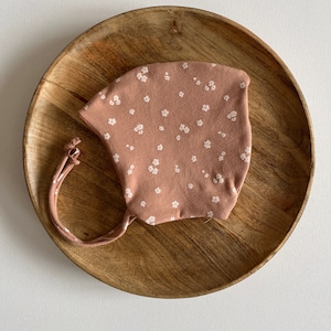 Op de afbeelding: Een roze stoffen mondkapje met een wit bloemenpatroon. Het masker heeft bandjes om het aan de kop te bevestigen.