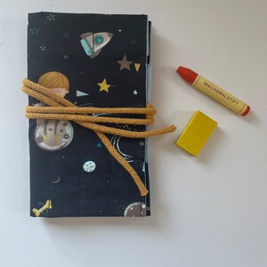 Op de afbeelding: Een zwarte stoffen oproletui met een ruimtevaartmotief van raketten, sterren en een figuur op een planeet, vastgezet met een mosterdgeel koord. Naast de etui liggen een oranje krijtje met de tekst WACHSMALSTIFT en een klein geel blokje.