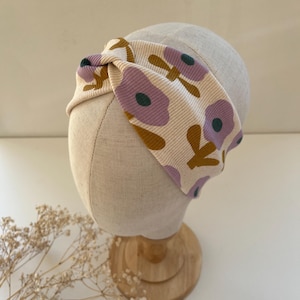 Könnte beinhalten: Ein cremefarbenes Blumenstirnband mit Knoten-Design. Das Stirnband hat einen gerippten Stoff mit einem sich wiederholenden Muster aus lila Blumen, grünen Zentren und braunen Blättern. Es wird auf einem Schaufensterkopf präsentiert.