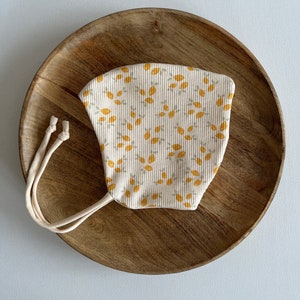 Op de afbeelding: Een wit katoenen gezichtsmasker met een gele citroenprint. Het masker heeft twee bandjes om het aan de kop te bevestigen.