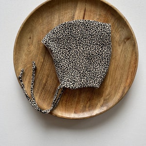 May include: A baby bonnet with a black and tan leopard print pattern. The bonnet is made of soft fabric and has tie closures. The bonnet is displayed on a wooden plate.