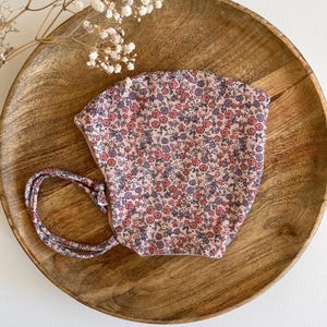 Pode incluir: Um gorro com estampa floral em tons de rosa, vermelho e roxo. O gorro apresenta um padrão delicado de pequenas flores e uma gravata a condizer. O gorro é colocado num prato de madeira.