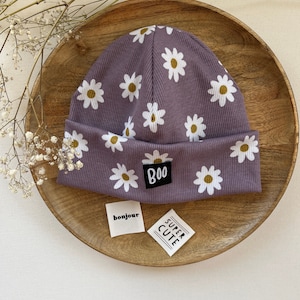 Puede incluir: Un gorro lila con un estampado de margaritas y un borde doblado. El gorro tiene margaritas blancas con centros amarillos. Una pequeña etiqueta negra con la palabra "BOO" está cosida en la parte delantera. También hay dos pequeñas etiquetas con las palabras "bonjour" y "SUPER CUTE".