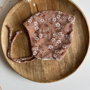 Pode incluir: Um gorro de bebé com um padrão floral. O gorro é de cor castanha suave com um padrão repetido de flores brancas e folhas castanhas. Tem laços em ambos os lados. O gorro está sobre um prato de madeira.