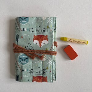 Op de afbeelding: Een stoffen etui met een vos- en bosontwerp in tinten groenblauw, oranje en bruin. De etui is vastgebonden met een bruin koord. Een gele krijt en een klein oranje blok liggen ernaast. Ongeveer 20 cm lang.