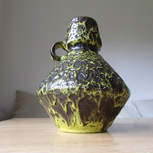 May include: A yellow and brown ceramic vase with a textured surface and a handle. The vase has a unique, abstract design.