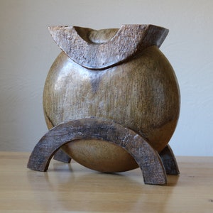 Könnte beinhalten: Eine runde, braune Keramikvase mit einem einzigartigen, geometrischen Design. Die Vase steht auf einem Holzständer mit zwei geschwungenen Stützen.