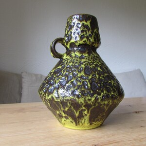 May include: A yellow and brown ceramic vase with a textured surface and a handle. The vase has a wide base and a narrow neck.