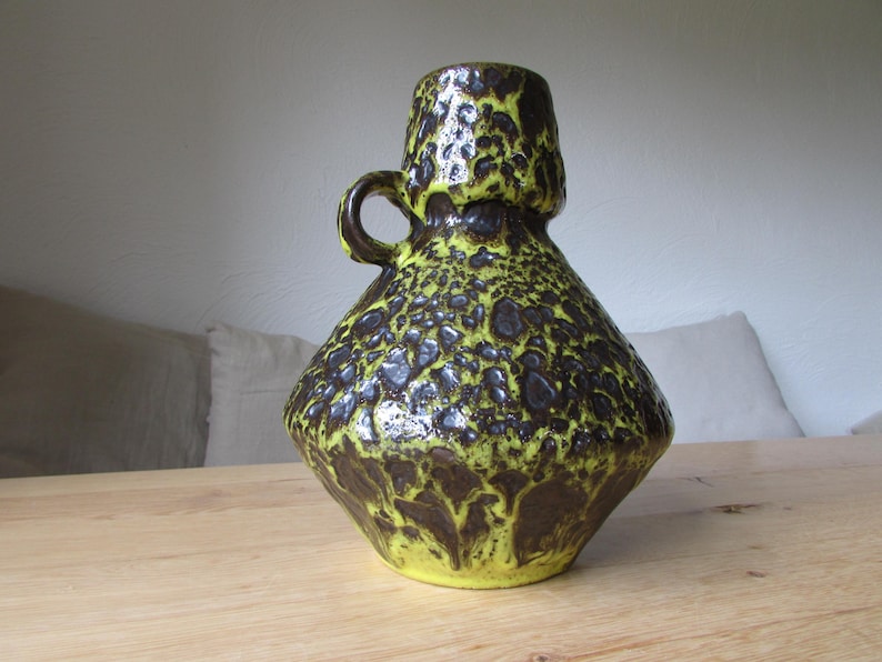 May include: A yellow and brown ceramic vase with a textured surface and a handle. The vase has a wide base and a narrow neck.