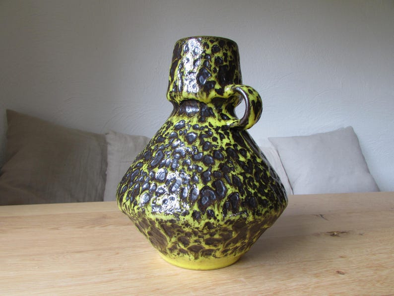 May include: A yellow and brown ceramic vase with a textured surface and a handle. The vase has a unique, abstract design.