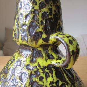May include: A yellow and brown ceramic vase with a textured surface and a handle. The vase has a unique, abstract design.