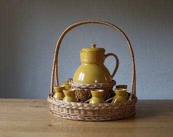 May include: A set of yellow ceramic serving pieces in a wicker basket. The set includes a jug and six cups.