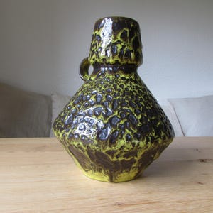 May include: A ceramic vase with a textured, mottled green and brown glaze. The vase has a wide, round base and a narrow neck with a handle.
