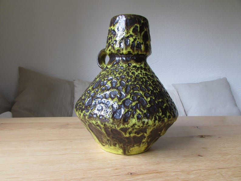 May include: A ceramic vase with a textured, mottled green and brown glaze. The vase has a wide, round base and a narrow neck with a handle.