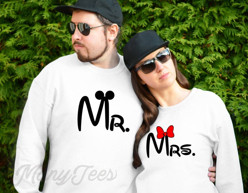 K&ouml;nnte beinhalten: Zwei wei&szlig;e Sweatshirts mit schwarzem Text und Grafiken. Der Text auf dem linken Sweatshirt lautet "Mr." mit Mickey-Maus-Ohren. Der Text auf dem rechten Sweatshirt lautet "Mrs." mit einer roten Schleife.