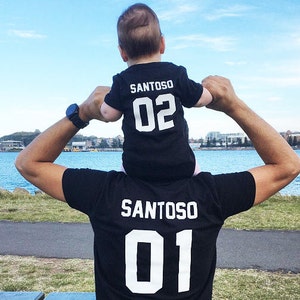 Pu&ograve; includere: Un padre e un figlio che indossano magliette nere abbinate con il nome "SANTOSO" e i numeri "01" e "02" stampati in bianco. Il padre tiene il figlio sulle sue spalle.