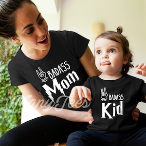 May include: A black t-shirt with white text that says "Badass Mom" and a black t-shirt with white text that says "Badass Kid". Both shirts feature a rock and roll hand gesture graphic.