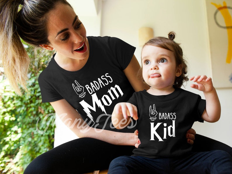 May include: A black t-shirt with white text that says "Badass Mom" and a black t-shirt with white text that says "Badass Kid". Both shirts feature a rock and roll hand gesture graphic.